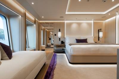 A luxurious bedroom with a plush white chaise lounge in the foreground and a large bed with ambient lighting in the background.