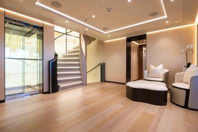 A modern, well-lit foyer with a glass elevator and a curved staircase in the foreground, and plush seating and a doorway in the background.