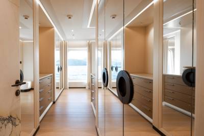 Mirrored closet doors reflect a hallway with wooden floors and drawers, leading to windows with a view of the ocean and distant hills.
