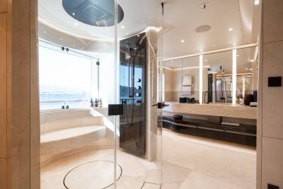 A modern, luxurious bathroom features a glass-enclosed shower with a curved bench in the foreground, and a vanity with a large mirror and ample lighting in the background.