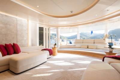 A luxurious cream-colored sectional sofa with red accent pillows and throws fills the foreground, while a panoramic view of a sun-drenched coastline with lush green hills is visible through large windows in the background.
