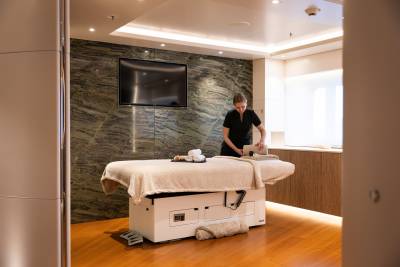 A massage therapist prepares a massage table in a spa room with a marble accent wall and a television in the background.