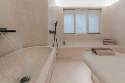 Water cascades into a modern bathtub in the foreground, while a massage table and a frosted window are visible in the background of this spa-like bathroom.