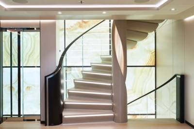 A modern, illuminated spiral staircase with a glass elevator to the left is set against a backdrop of a large, backlit agate-patterned wall.