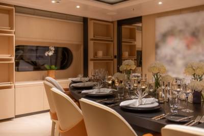 A formal dining table set with white napkins and floral arrangements is in the foreground, while a wall with shelves and a large abstract painting is in the background.