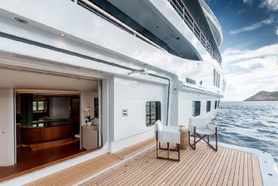 Two chairs and a small table sit on the wooden deck of a large white yacht, with the ocean and a distant island visible in the background.