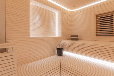 A modern wooden sauna with a bucket and towels in the foreground and illuminated slatted walls and benches in the background.