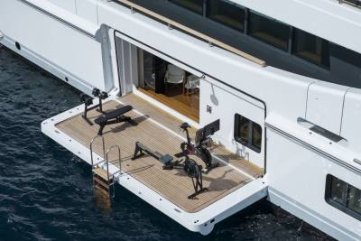 A modern yacht's open aft deck features a well-equipped gym with rowing machines and a weight bench, while the calm blue water surrounds the vessel.