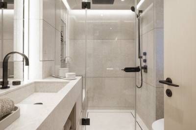 A modern bathroom features a sleek vanity with black faucets in the foreground and a glass-enclosed shower with tiled walls in the background.