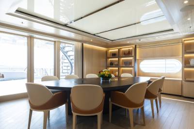 A large oval dining table with chairs is in the foreground, while a bright, modern room with shelves and large windows is in the background.