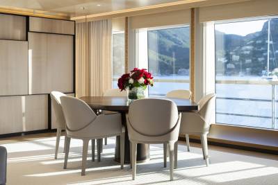 A dining table with a floral centerpiece is set in a room with large windows overlooking a body of water and a mountainous landscape.