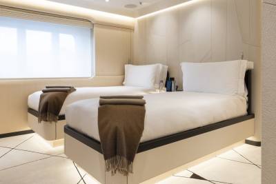 Two neatly made twin beds with brown throws are in the foreground, while a large window with blinds and a textured wall are in the background.