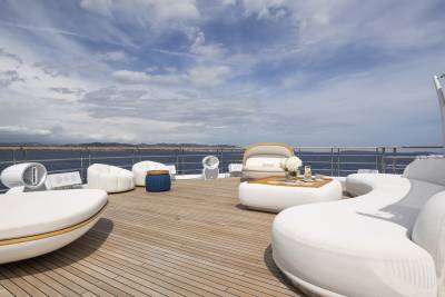 Luxurious white outdoor furniture is arranged on a wooden deck with a cloudy blue sky and distant coastline in the background.