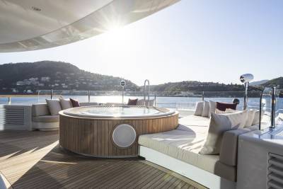 A circular jacuzzi and plush seating on a yacht's deck are bathed in sunlight, with a coastal town and hills visible in the background.