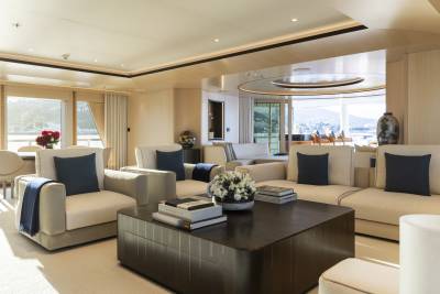 A spacious, modern living area with plush white sofas and a dark wood coffee table in the foreground, and large windows revealing a sunlit outdoor dining area and a distant coastal view in the background.