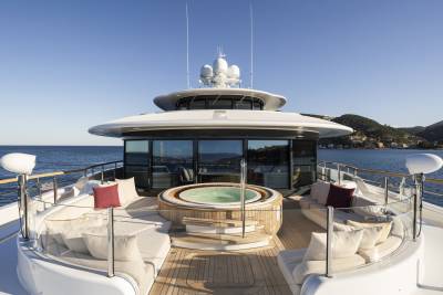 A luxurious yacht deck with a hot tub and seating in the foreground overlooks a calm sea and a distant, tree-covered coastline under a clear blue sky.