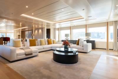 A spacious, modern living area with a large white sectional sofa and a dark coffee table in the foreground, and large windows overlooking a waterfront view in the background.