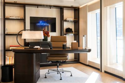 A modern office with a brown leather desk and chair in the foreground, and built-in shelves with a television and windows in the background.