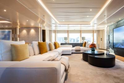 A luxurious white sectional sofa with colorful throw pillows sits in the foreground of a spacious yacht lounge, while a large television screen displays a scenic mountain landscape in the background.