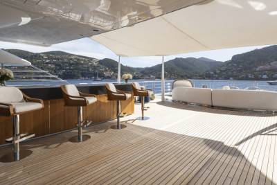 A luxurious yacht deck with a polished wooden floor, a sleek bar with stools in the foreground, and a scenic coastal view with mountains and other boats in the background.