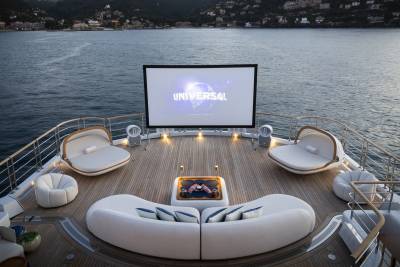 A large outdoor movie screen is set up on the deck of a yacht, with comfortable seating arranged around a fire pit, all set against a backdrop of a calm sea and a distant coastline.