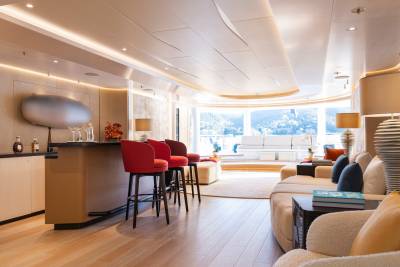 A modern yacht interior features a sleek bar with red stools in the foreground and a spacious lounge area with large windows overlooking a scenic landscape in the background.