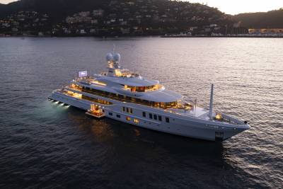 A large, illuminated yacht floats on calm water in the foreground, with a hillside town and mountains visible in the dusky background.
