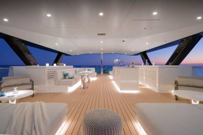 White lounge chairs and a pouf are in the foreground of a modern yacht deck with a bar and seating area, overlooking the ocean at dusk.