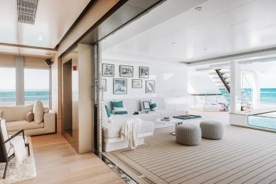 A spacious, modern yacht interior with a white sectional sofa and ottomans in the foreground, and a bright, open living area with a view of the ocean in the background.