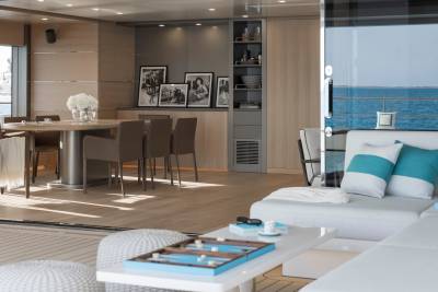 A modern yacht interior features a living area with a backgammon board in the foreground and a dining area with framed photos on the wall in the background, overlooking the ocean.