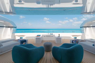 Two plush teal armchairs face a serene ocean view from a modern yacht deck, with white lounge chairs and a gray ottoman in the foreground and a bright blue sky with scattered clouds in the background.