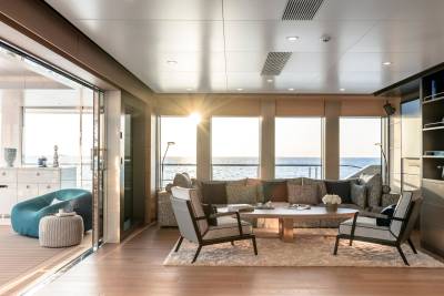 Sunlight streams into a luxurious yacht lounge with comfortable seating in the foreground and a serene ocean view through large windows in the background.