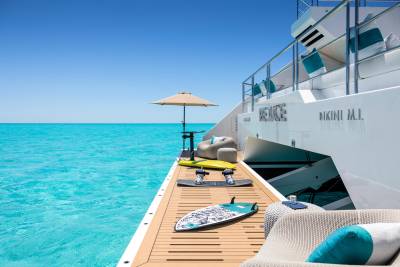 Wakeboards and a lounge chair sit on the deck of a yacht anchored in bright turquoise water under a clear blue sky.