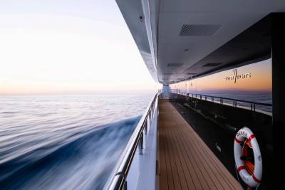 A wooden deck with a life preserver runs along the side of a yacht as it cruises through calm ocean waters under a soft, hazy sky.