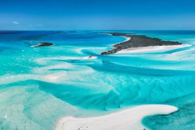 Shallow, turquoise waters with sandbars in the foreground give way to a distant, lush green island under a clear blue sky.