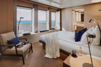 A luxurious yacht cabin with a comfortable armchair in the foreground and a large bed facing windows with a view of the ocean in the background.