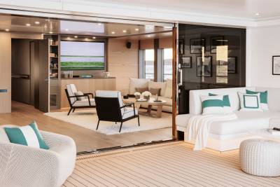 A modern yacht's living area features a white sectional sofa with teal-accented pillows in the foreground, opening to a lounge with armchairs and a television in the background.