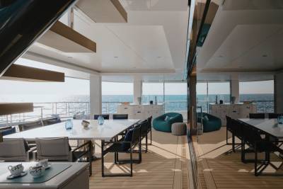 Modern outdoor dining areas with white tables and chairs are set against a backdrop of the calm ocean and a bright sky, reflected in a mirrored wall.