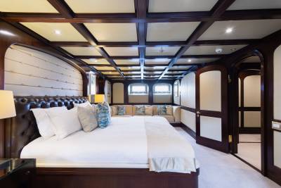 A luxurious bedroom with a tufted headboard and plush bedding in the foreground, and a seating area with windows and an adjoining hallway in the background.