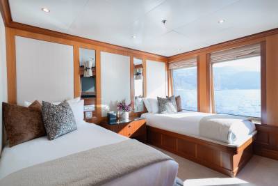 Two beds with plush pillows and blankets fill the foreground, while a large window in the background reveals a serene body of water and distant mountains.