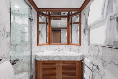 A luxurious bathroom features a marble vanity with a tri-fold mirror in the foreground, while the background reveals a glass-enclosed shower and marble-clad walls.