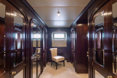 A plush chair sits in the center of a polished wood hallway with mirrored doors on either side, leading to a window with blinds in the background.