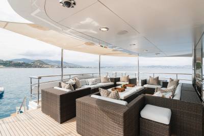 A spacious, covered outdoor lounge area with wicker furniture and plush cushions sits on a yacht's deck, overlooking a calm sea and a distant, hilly coastline.
