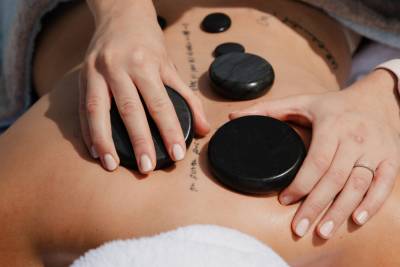Hands place smooth, dark stones on a tattooed back, with a white towel visible in the foreground and background.