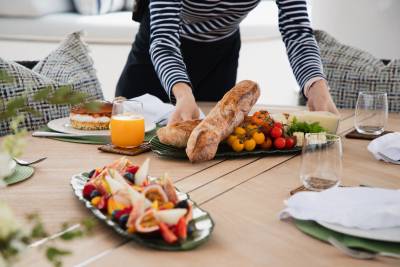 A person in a striped shirt sets a table with a fruit salad, bread, tomatoes, and cheese on a yacht deck, with cushioned seating visible in the background.