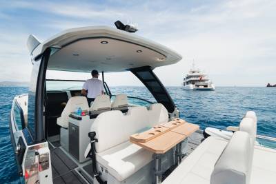 A man steers a modern speedboat with plush seating and a fold-out table in the foreground, while a large white yacht sails in the background under a clear blue sky.