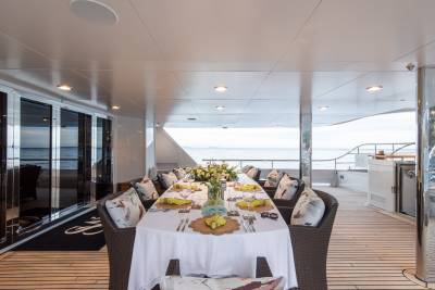 A long dining table set for a meal on a yacht's deck is in the foreground, with the ocean and sky visible in the background.