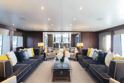 A luxurious yacht lounge with plush sofas and armchairs in the foreground, and a dining area visible through glass doors in the background.