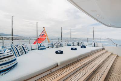 A spacious, white cushioned deck with blue and white striped pillows and a British flag in the foreground overlooks a calm sea and distant coastline in the background.