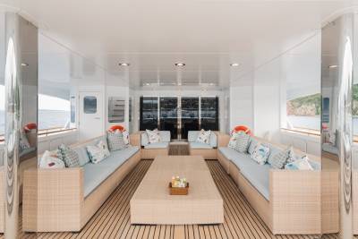 A spacious, light-filled yacht deck with comfortable seating and a coffee table in the foreground, and a scenic ocean view with distant islands in the background.